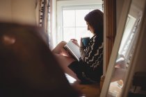 Donna seduta vicino alla finestra e che legge un libro a casa — Foto stock