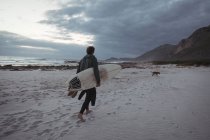 Hombre llevando tabla de surf caminando en la playa al atardecer - foto de stock