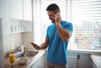 Man talking on mobile phone while using digital tablet in kitchen at home — Stock Photo