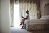 Donna anziana seduta sul letto e libro di lettura in camera da letto — Foto stock