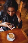 Hermosa mujer haciendo clic en la foto de café desde el teléfono móvil en la cafetería - foto de stock