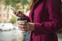 Средняя секция бизнесвумен держит одноразовый кофейный стакан и с помощью мобильного телефона на улице — стоковое фото