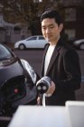 Улыбающийся мужчина держит зарядное устройство для автомобиля на электростанции зарядки автомобиля — стоковое фото