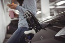 Auto in carica con caricabatterie elettrico mentre la donna in piedi in background presso la stazione di ricarica del veicolo elettrico — Foto stock