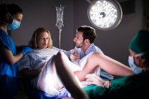 Доктор слідчим вагітної жінки під час доставки під час чоловік тримає руку в операційний зал — стокове фото