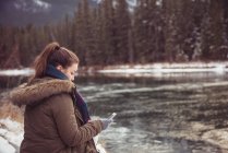Жінка сидить на березі річки і використовує мобільний телефон взимку — стокове фото