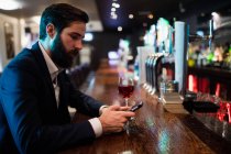 Uomo d'affari che utilizza il telefono cellulare con bicchiere di vino sul bancone nel bar — Foto stock