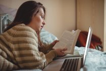 Mulher bonita lendo um livro e usando laptop na cama em casa — Fotografia de Stock
