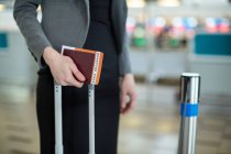 Parte média da empresária de pé com cartão de embarque, passaporte e bagagem no terminal do aeroporto — Fotografia de Stock