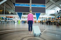 Rear view of female commuter standing with luggage at waiting area in airport — Stock Photo