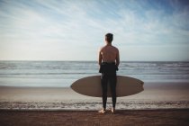 Vista trasera del surfista de pie con tabla de surf en la playa - foto de stock