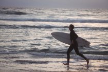 Vista lateral de un hombre llevando tabla de surf corriendo en la playa al atardecer - foto de stock