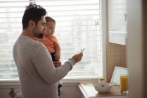 Mid adult man using mobile phone while holding baby son in kitchen at home — Stock Photo