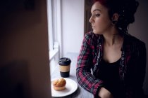 Mujer reflexiva mirando a través de la ventana con cupcake y taza de café en la mesa en la cafetería - foto de stock