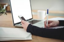 Geschäftsführender Angestellter schreibt Tagebuch, während er sein Handy im Büro benutzt — Stockfoto