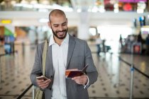 Homme d'affaires souriant tenant une carte d'embarquement et vérifiant son téléphone portable au terminal de l'aéroport — Photo de stock