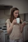 Frau trinkt Kaffee, während sie ihr Handy zu Hause in der Küche benutzt — Stockfoto
