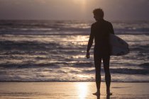 Silhueta de um homem carregando prancha de surf em pé na praia ao entardecer — Fotografia de Stock