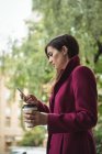 Donna d'affari in possesso di tazza di caffè usa e getta e utilizzando il telefono cellulare sulla strada — Foto stock
