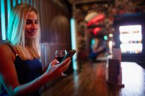 Mulher bonita usando telefone celular, tendo vinho tinto no balcão no bar — Fotografia de Stock