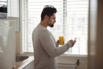Uomo che usa il telefono cellulare mentre ha succo in cucina a casa — Foto stock