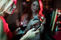 Mulher bonita usando telefone celular, tendo vinho tinto no balcão no bar — Fotografia de Stock