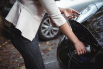 Mid section of woman using mobile phone while charging electric car on street — Stock Photo