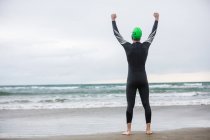 Vue arrière d'un athlète heureux debout sur la plage les mains levées — Photo de stock