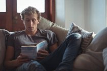 Uomo seduto sul divano a leggere un libro in salotto — Foto stock