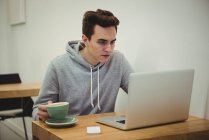 Человек смотрит на ноутбук, держа чашку кофе в кофейне — стоковое фото