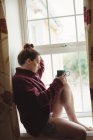 Nachdenkliche Frau sitzt auf Fensterbank und hält Kaffeetasse zu Hause — Stockfoto