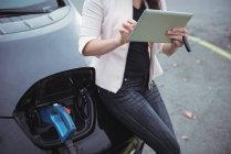 Partie médiane de la femme utilisant une tablette numérique tout en rechargeant la voiture électrique sur la rue — Photo de stock