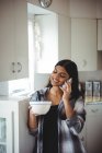 Mulher falando no celular enquanto toma café da manhã na cozinha — Fotografia de Stock