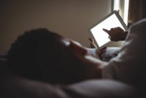 Rückansicht eines Mannes, der sein digitales Tablet benutzt, während er sich im Schlafzimmer auf dem Bett entspannt — Stockfoto