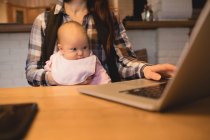 Mãe carregando bebê enquanto usa laptop na mesa em casa — Fotografia de Stock