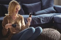 Красива жінка використовує мобільний телефон, маючи чай у вітальні вдома — стокове фото