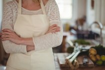 Sección media de la mujer de pie con los brazos cruzados en la cocina en casa - foto de stock