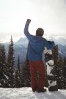 Rear view of man with hand raised holding snowboard on mountain against sky during winter — Stock Photo