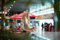 Mulher sorridente com café em pé na área de espera no terminal do aeroporto — Fotografia de Stock