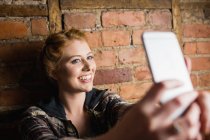 Femme debout contre un mur de briques et prenant un selfie sur son téléphone portable — Photo de stock