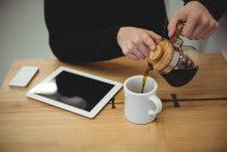 Sección media del hombre vertiendo café de la tetera en la taza en la cafetería - foto de stock