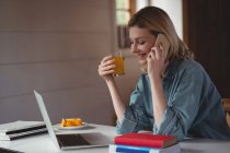 Beautiful woman talking on mobile phone while having juice at home — Stock Photo