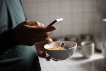 Meio da seção de homem usando telefone celular enquanto toma café da manhã na cozinha em casa — Fotografia de Stock