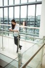 Бізнес-леді, спираючись на перила і використовуючи мобільний телефон в офісному коридорі — стокове фото