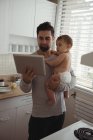 Чоловік середнього віку використовує цифровий планшет, тримаючи дитину на кухні — стокове фото