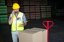Joven trabajador masculino sujetando el portapapeles mientras habla por teléfono en fábrica - foto de stock