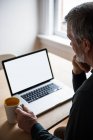 Homem olhando para laptop enquanto toma uma xícara de café em casa — Fotografia de Stock
