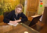 Femme assise au bureau avec un ordinateur portable et écrivant sur un bloc-notes dans le bureau — Photo de stock