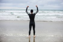 Rear view of athlete in wet suit standing with arms up on beach — Stock Photo