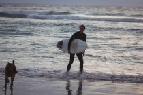 Homme portant une planche de surf debout sur la plage avec son chien au crépuscule — Photo de stock